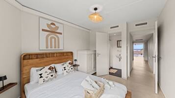 A hotel room with a wooden bed, a bedside table, a lamp, a wall-mounted artwork, a hanging light fixture, and a view of the sea through an open door.