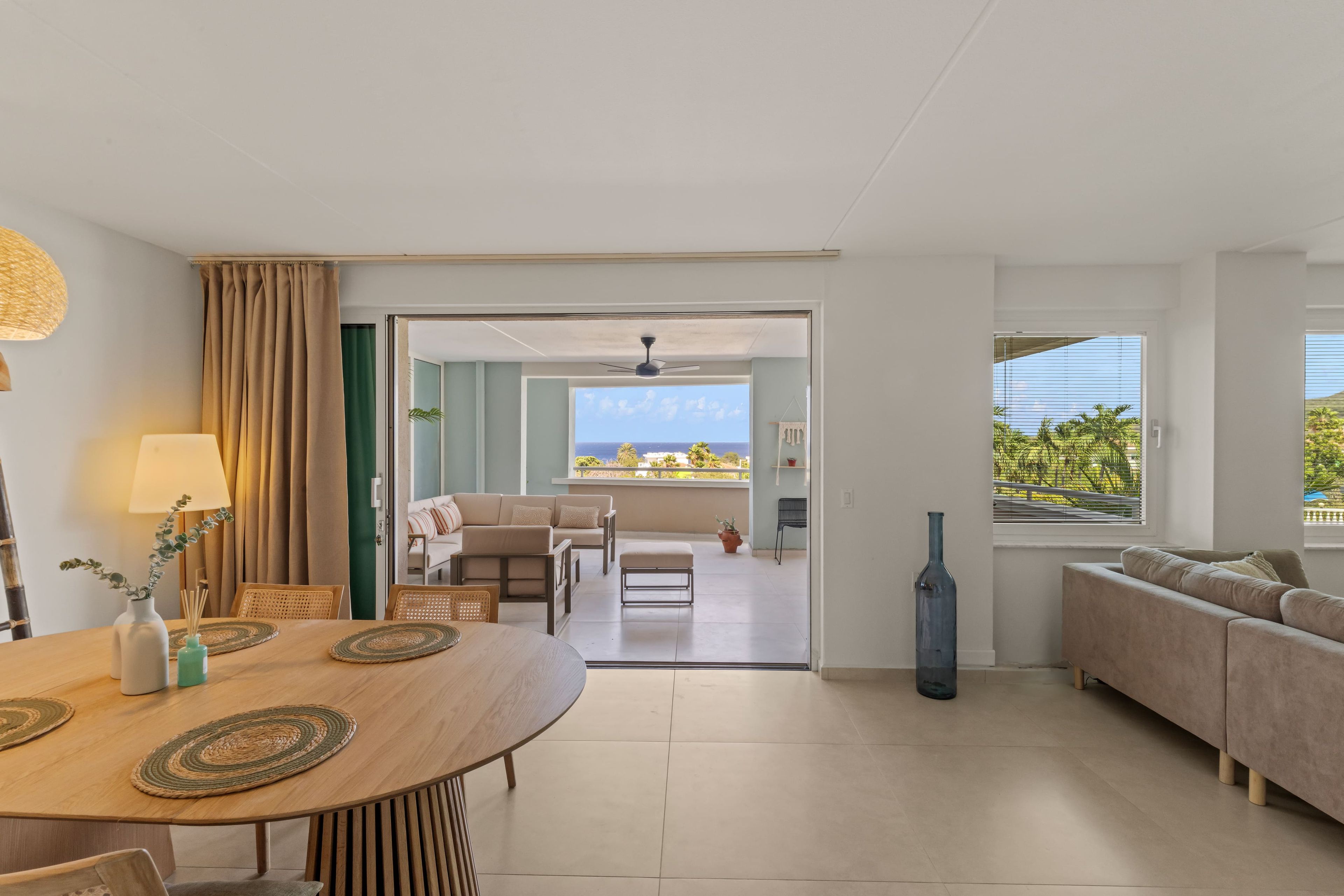 A modern living room with a dining area, a sofa, a table with a lamp, and a view of the outdoors.