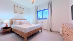 A bedroom with a wooden bed, a nightstand with a lamp, a dresser, a window with curtains, and a wall-mounted picture. - Serene Pool View Walk to Fun Beach Dine Shop (Punta Cana)
