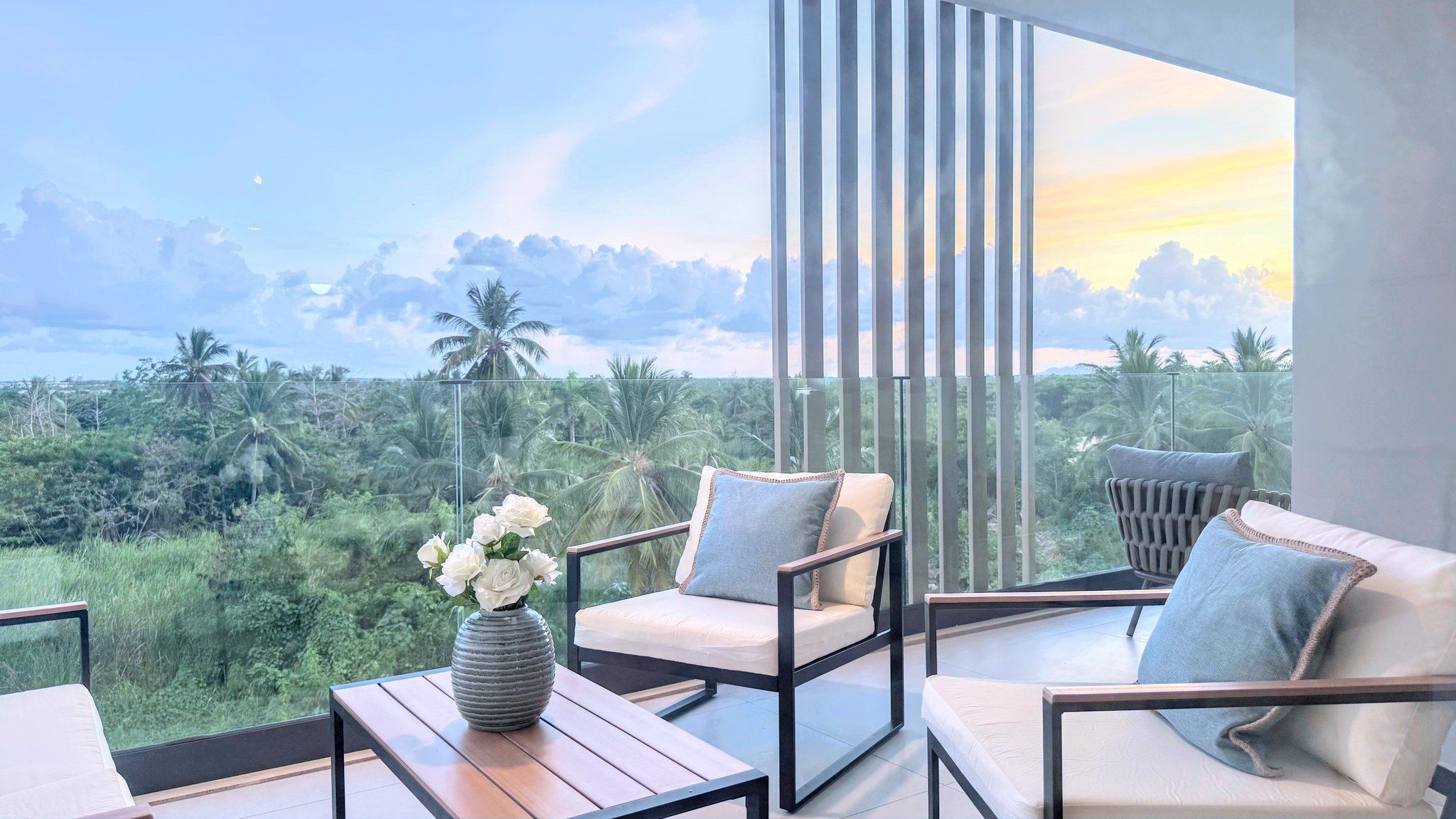 Ein Balkon mit Blick auf eine tropische Landschaft, mit einem Tisch, einer Blumenvase, zwei Stühlen und einem Sofa.