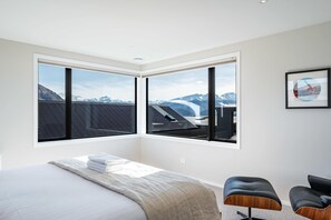 A bedroom with a large bed, two black chairs, and a view of mountains through large windows. - Eliza's Escape with Mountain Views (Wānaka)