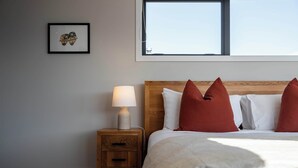 Un lit bien fait, avec une literie blanche et des oreillers rouges, une lampe de chevet, une table de chevet en bois avec un tiroir et un tableau encadré au mur.