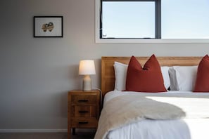 A neatly made bed with white bedding and red pillows, a bedside lamp, a wooden nightstand with a drawer, and a framed picture on the wall. - Eliza's Escape with Mountain Views (Wānaka)