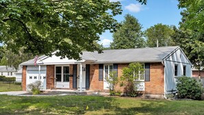 A single-story brick house with a front porch, a flag, and a small front yard. - Oklahoma Breeze | Walk to Track & Downtown | 4BR + Pet Friendly (Saratoga Springs)