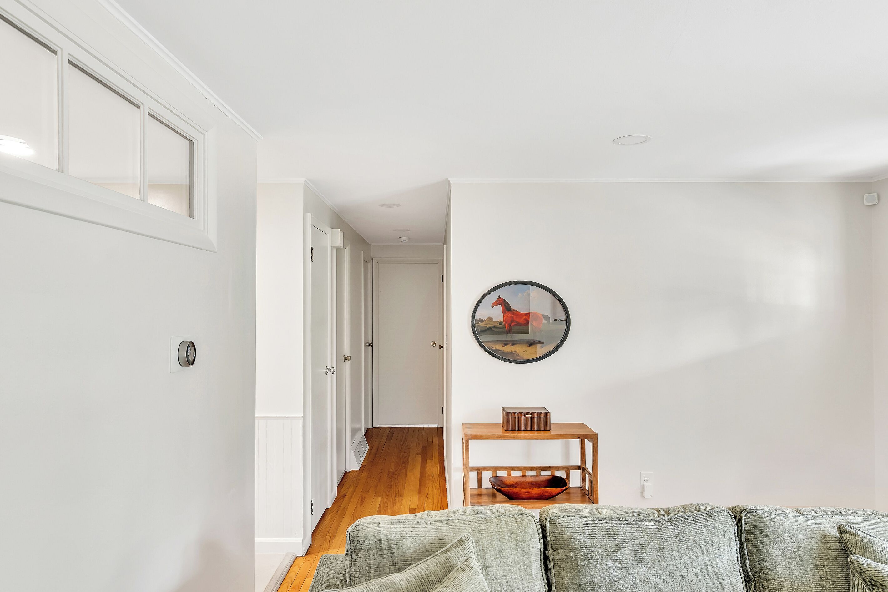 A modern living room with a wooden floor, a grey sofa, a wooden side table with a bowl, and a door leading to another room.