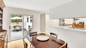 A modern kitchen with white cabinetry, a wooden dining table, and a view of an outdoor patio. - Oklahoma Breeze | Walk to Track & Downtown | 4BR + Pet Friendly (Saratoga Springs)