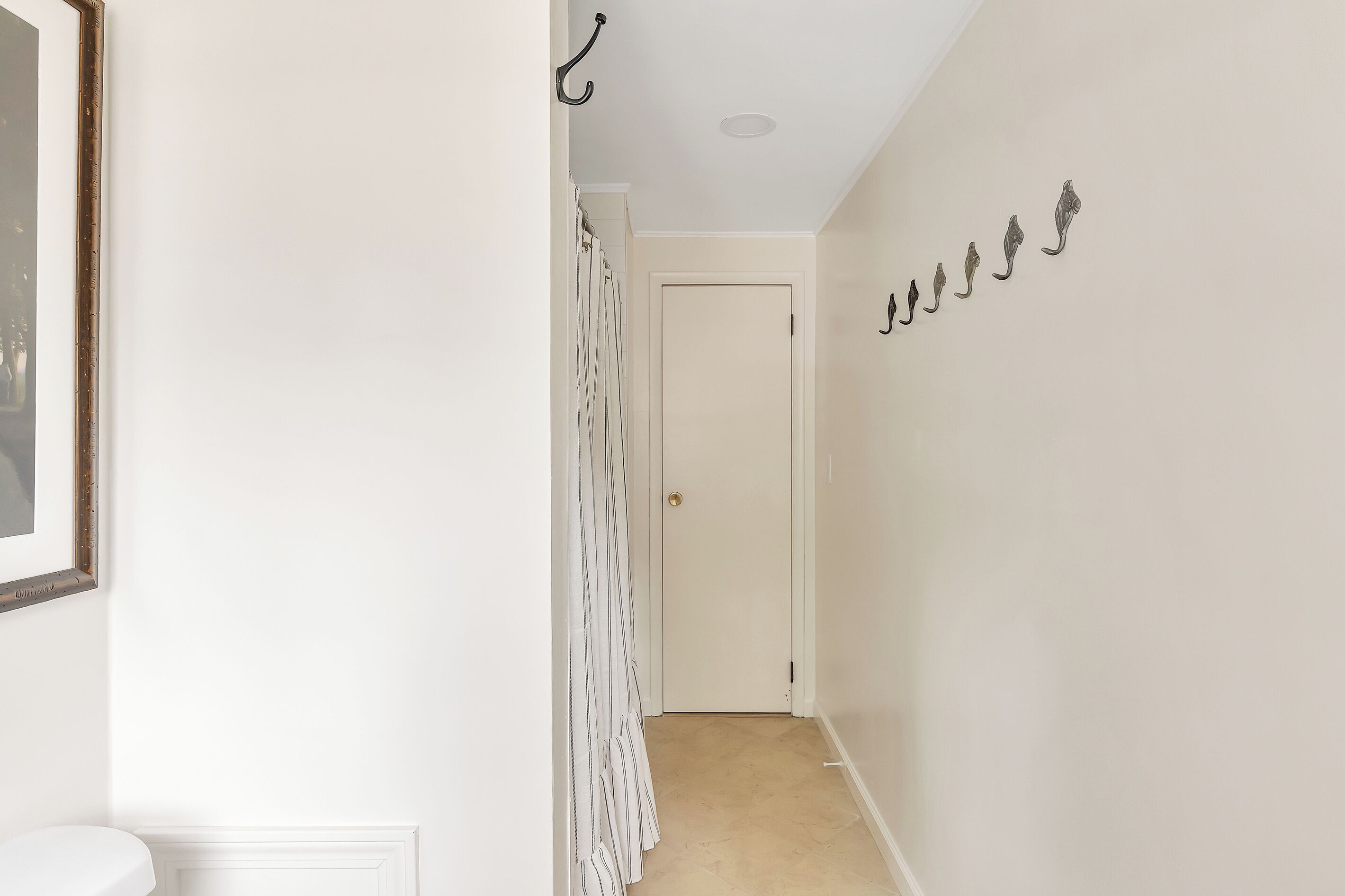 A hallway with a white door at the end, a coat rack with several coats, and a framed picture on the wall.