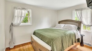 A bedroom with a bed, a dresser, a nightstand, and a window with curtains. - Oklahoma Breeze | Walk to Track & Downtown | 4BR + Pet Friendly (Saratoga Springs)