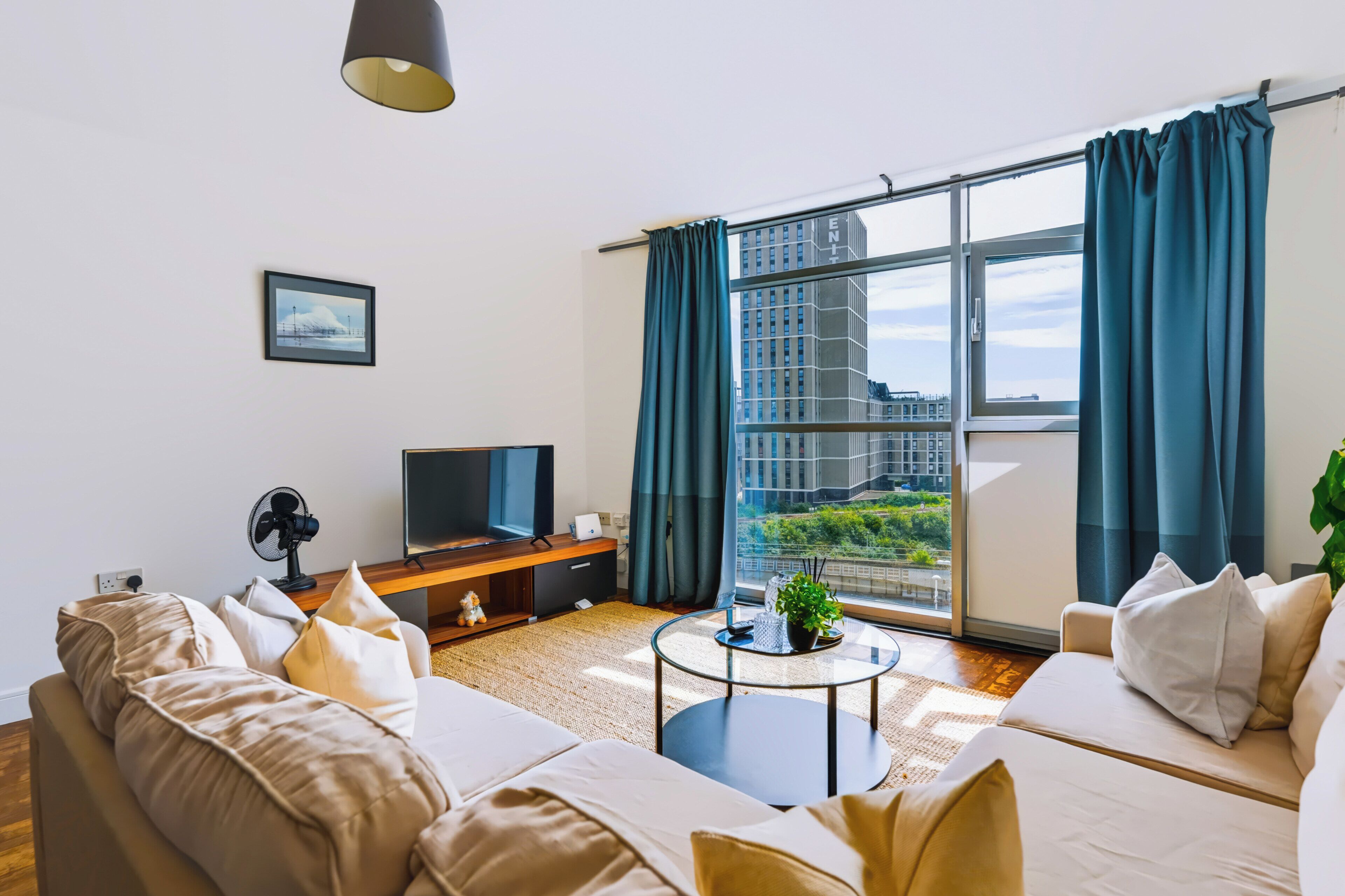 A modern living room with a sofa, a television, a small table, and a large window with curtains.