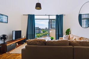 A modern living room with a large window, a sofa, a TV, and a fan. - Spacious 2-Bed 2-Bath Cardiff City Centre Apt (Cardiff)