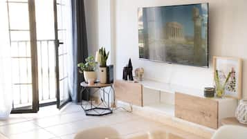 Ein modernes Wohnzimmer mit einem großen Flachbildfernseher an der Wand, einem runden Beistelltisch mit Dekoration, einer Bank mit einer kleinen Statue und Blick auf ein klassisches Gebäude durch das Fenster.