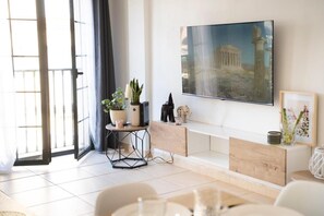 A modern living room with a large flat-screen TV mounted on the wall, a round side table with decorative items, a bench with a small statue, and a view of a classical building through the window. - Apartamento 'Estrella Del Sur Torremolinos' con vistas a la montaña, Wi-Fi y aire acondicionado (Torremolinos)
