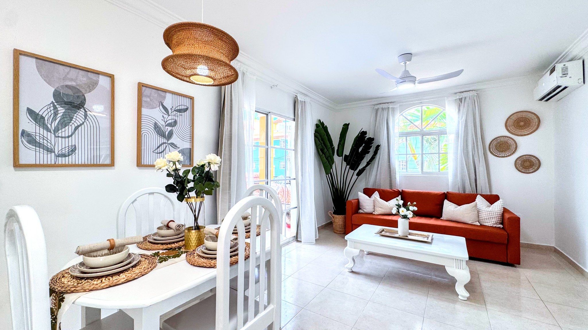 A well-lit living room with a white sofa, a dining table with a vase of flowers, and artwork on the walls.