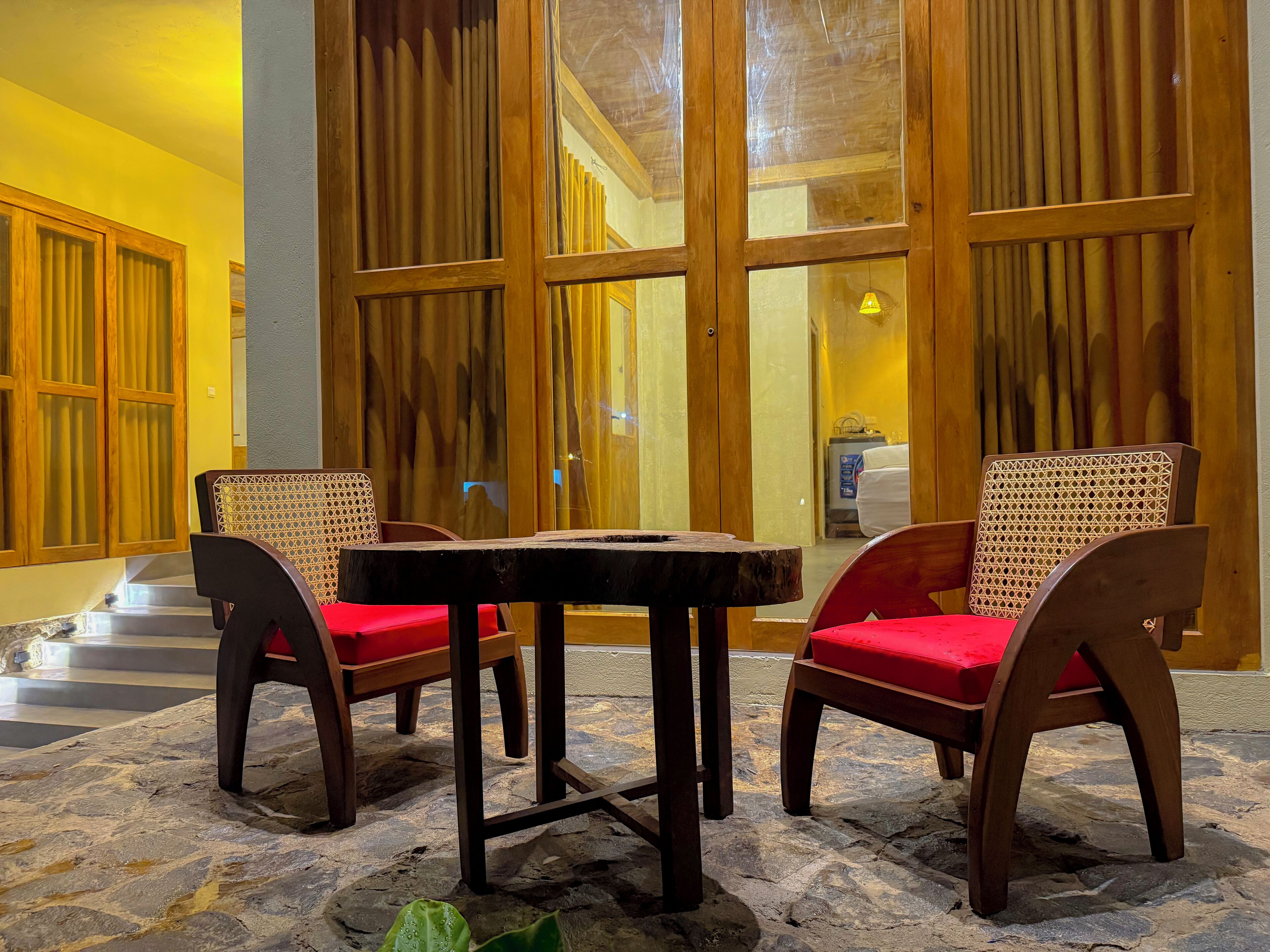 A stone-floored outdoor seating area with wooden furniture and red cushions, a wooden door with glass panels, and a yellow wall.