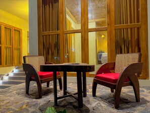 A stone-floored outdoor seating area with wooden furniture and red cushions, a wooden door with glass panels, and a yellow wall.