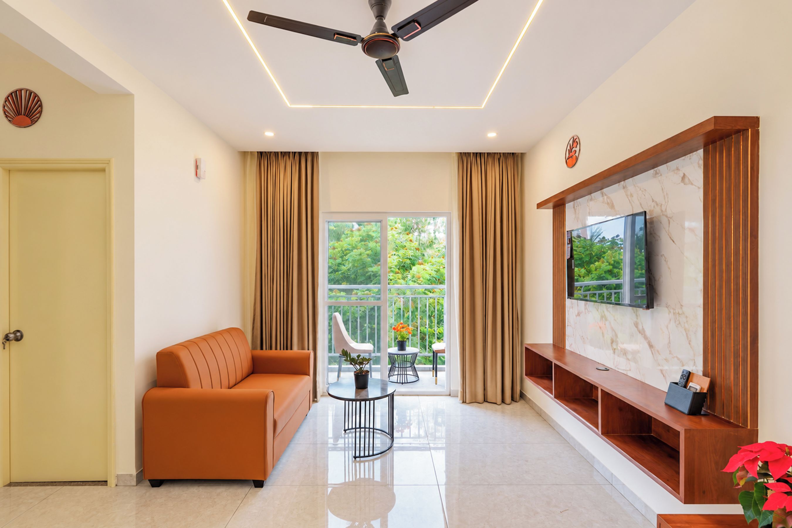 Uma sala de estar moderna com ventilador de teto, sofá, TV em parede de mármore e varanda com vista para o verde.