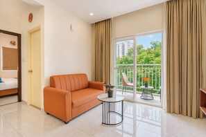 A modern hotel room with a sofa, a small round table, and a balcony with a view of greenery and buildings. - Terra Rose Suites By Bestinn Leisure (Bengaluru)