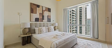 Um quarto moderno com uma cama grande, uma mesinha de cabeceira, uma luminária, uma obra de arte na parede e uma janela com vista para edifícios altos.