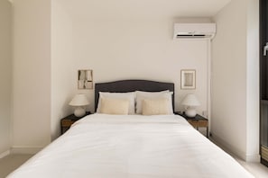 A hotel room with a bed, two bedside tables with lamps, an air conditioning unit, and two framed pictures on the wall. - Terrace Light in the Vale (Greater London)
