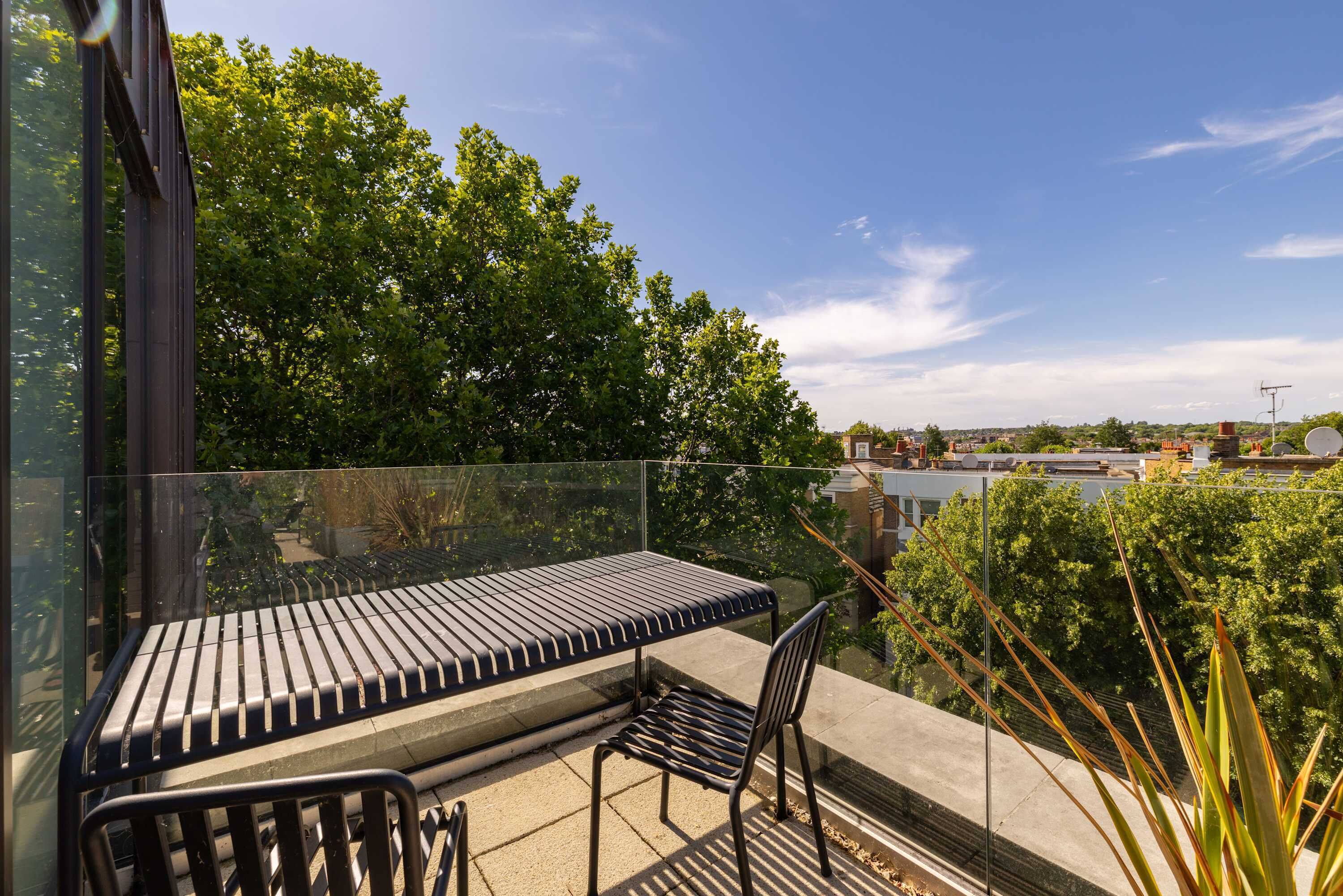 A balcony with a metal bench and chair, surrounded by glass railings, overlooking a cityscape with trees and buildings.