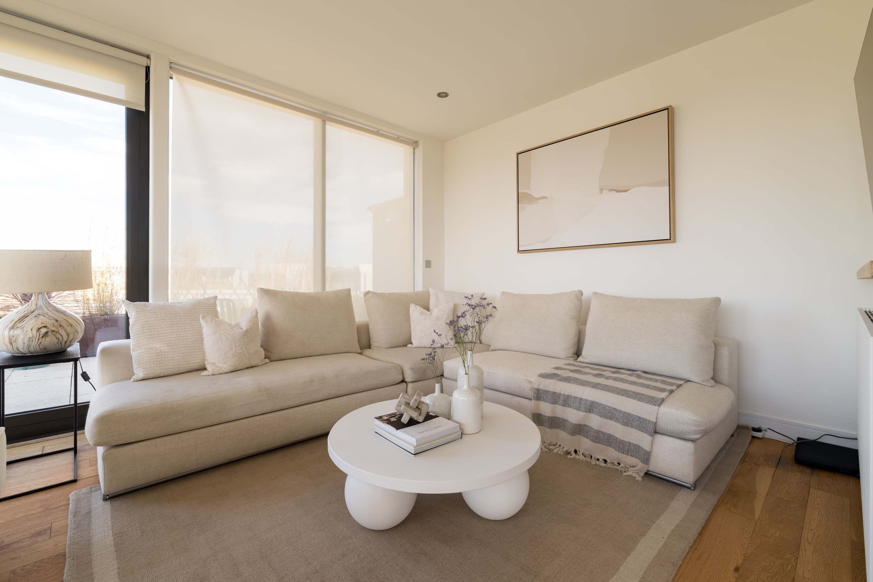 A modern living room with a beige sectional sofa, a white coffee table, and a large window providing natural light.