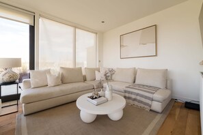 A modern living room with a beige sectional sofa, a white coffee table, and a large window providing natural light. - Terrace Light in the Vale (Greater London)