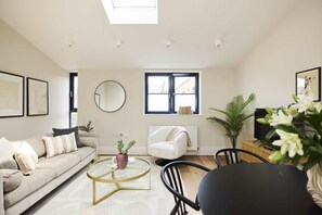 A modern living room with a sofa, coffee table, and a chair. There is a window with blinds, a radiator, and a television. - A Place to Dwell - Tooting (Greater London)
