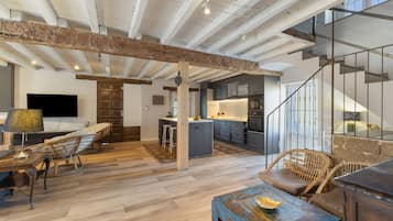 Amplia sala de estar con vigas de madera a la vista, cocina con electrodomésticos modernos y una escalera que conduce a un nivel superior.