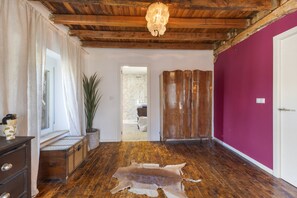 A room with wooden flooring, a hanging light fixture, a large mirror, a potted plant, and a door leading to another room.