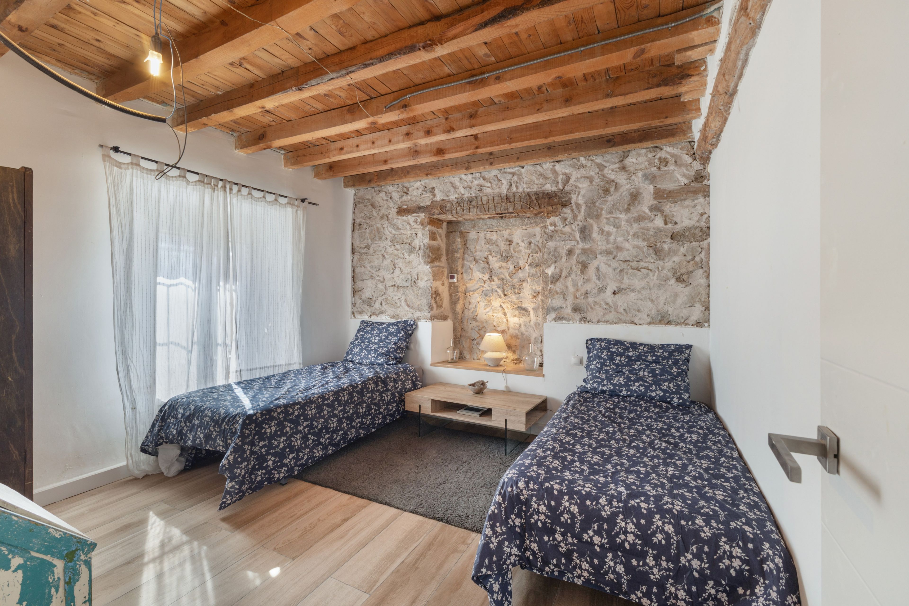 A bedroom with two single beds, a stone wall, a wooden ceiling, and a window with curtains.