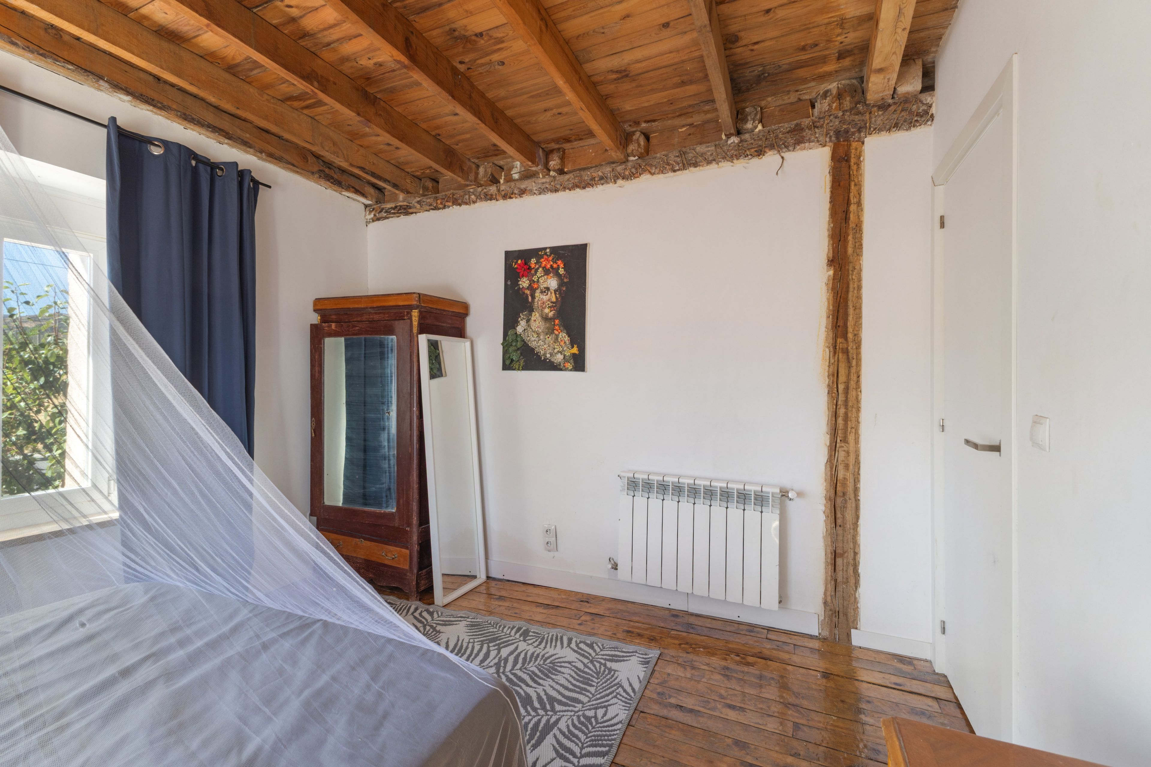 A room with a wooden ceiling, a bed with a white bedspread, a radiator, and a painting on the wall.