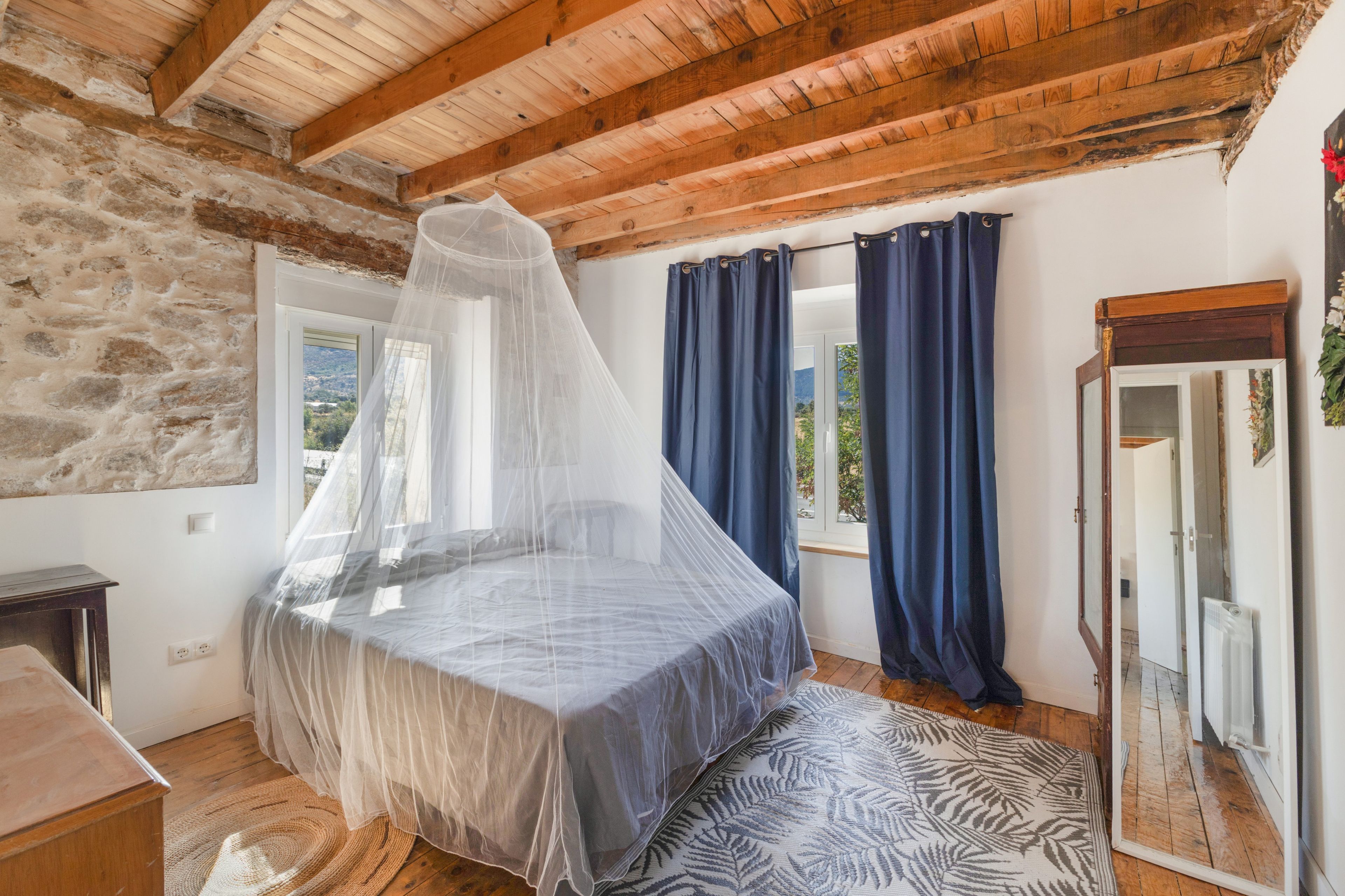 A bedroom with a bed under a mosquito net, wooden beams on the ceiling, a stone wall, a window with curtains, and a mirror.