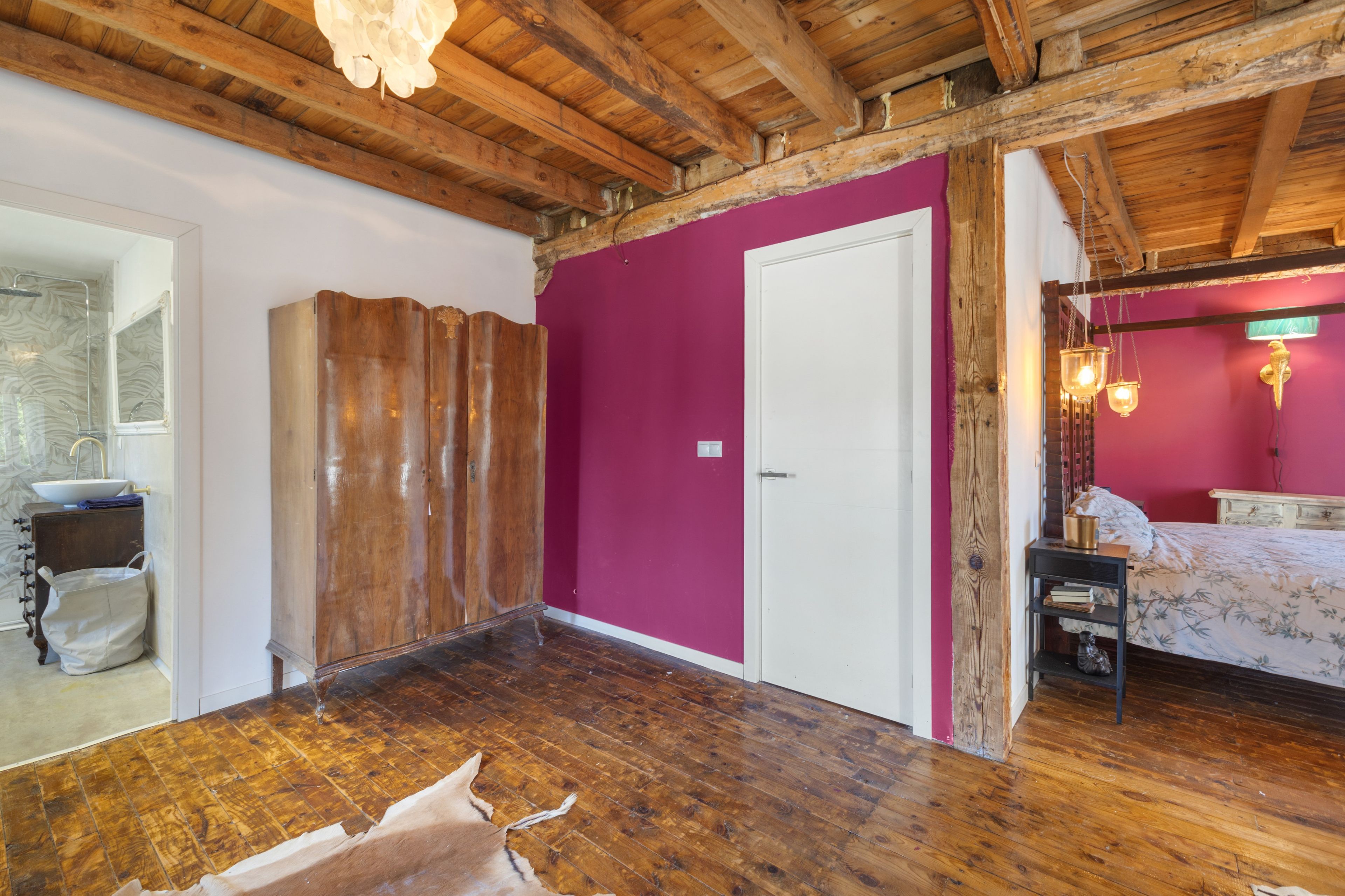Habitación con vigas de madera, un armario de madera, una puerta blanca y una cama con colcha estampada.