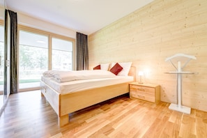 A bedroom with a wooden bed, a nightstand, a lamp, and a view through a large window. - Gartenferienwohnung Alvier Lodge (Brand)