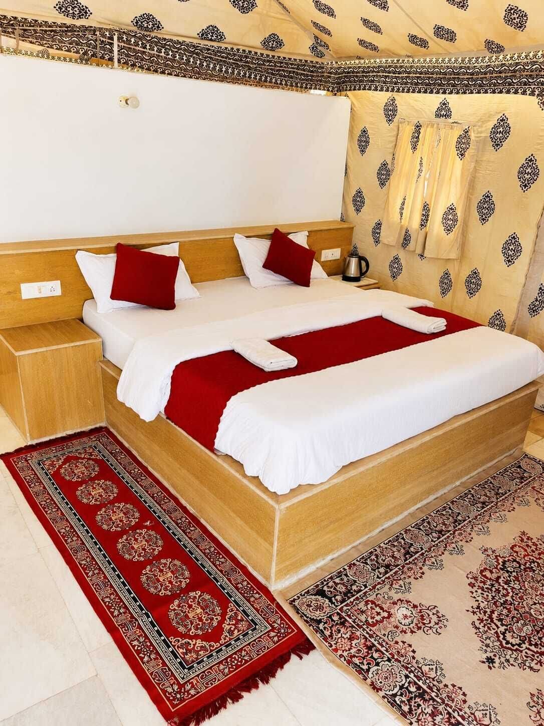 A hotel room with a wooden bed, white bedding, red pillows, a red and beige patterned rug, and a patterned wall.