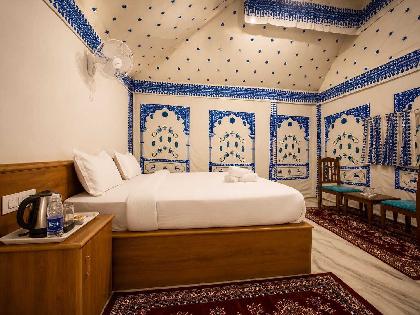 A hotel room with a bed, a wooden nightstand, a kettle, a bottle of water, and a table with a chair.