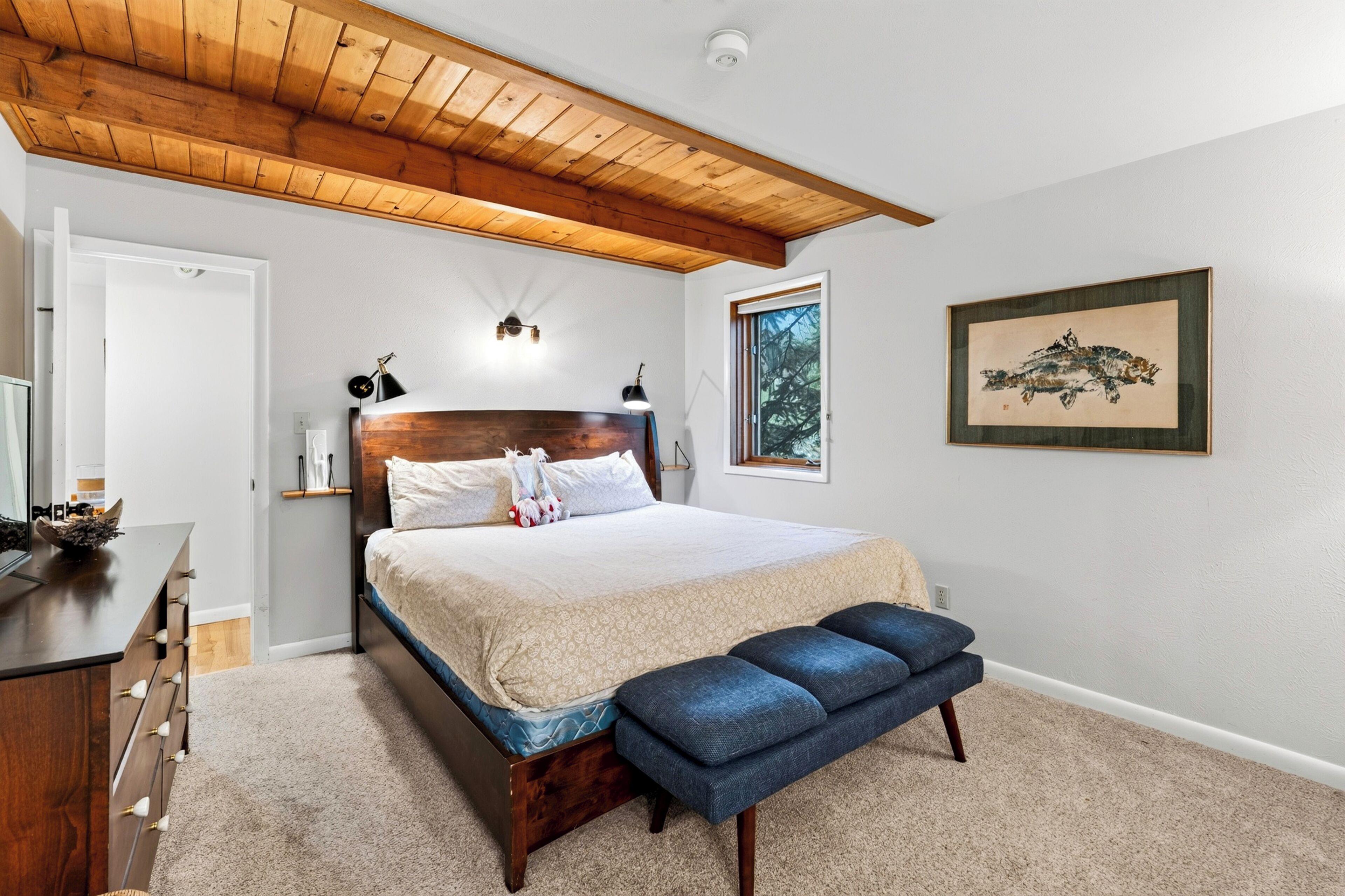 A bedroom with a wooden ceiling, a bed with a headboard, bedside lamps, a bench, a dresser, and a framed picture on the wall.