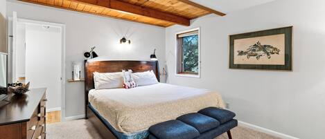 A bedroom with a wooden ceiling, a bed with a headboard, bedside lamps, a bench, a dresser, and a framed picture on the wall.