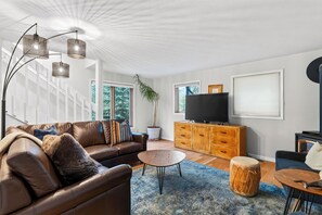 A modern living room with a leather sofa, a coffee table, a TV on a wooden cabinet, and a large window with a view of trees. - A-Frame + Hot Tub + Arcade + BBQ | Forest Views + Ski Access | Pitch & Pine (Breckenridge)