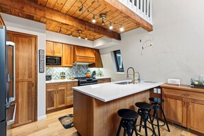 A modern kitchen with wooden cabinets, a large island with stools, a microwave, and a stove. - A-Frame + Hot Tub + Arcade + BBQ | Forest Views + Ski Access | Pitch & Pine (Breckenridge)