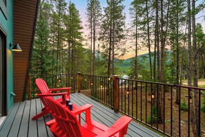 A wooden deck with red chairs and a table, overlooking a forested area with a view of distant hills. - A-Frame + Hot Tub + Arcade + BBQ | Forest Views + Ski Access | Pitch & Pine (Breckenridge)