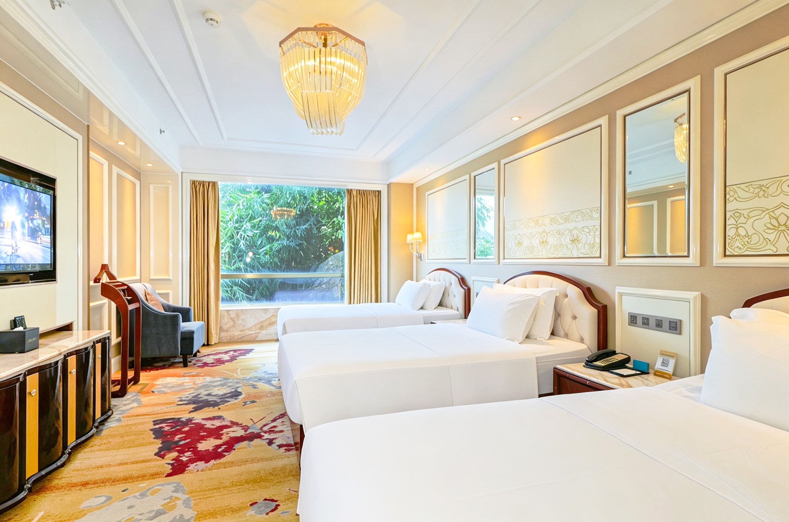 A hotel room with two beds, a TV, a desk, and a view of greenery.