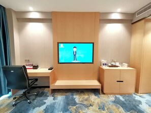 A hotel room with a flat-screen TV mounted on the wall, a desk with a chair, a cabinet, and a patterned carpet.