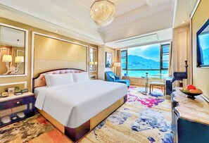 A hotel room with a large bed, a desk with a chair, a dining table with chairs, a large window with a mountain view, and a chandelier.