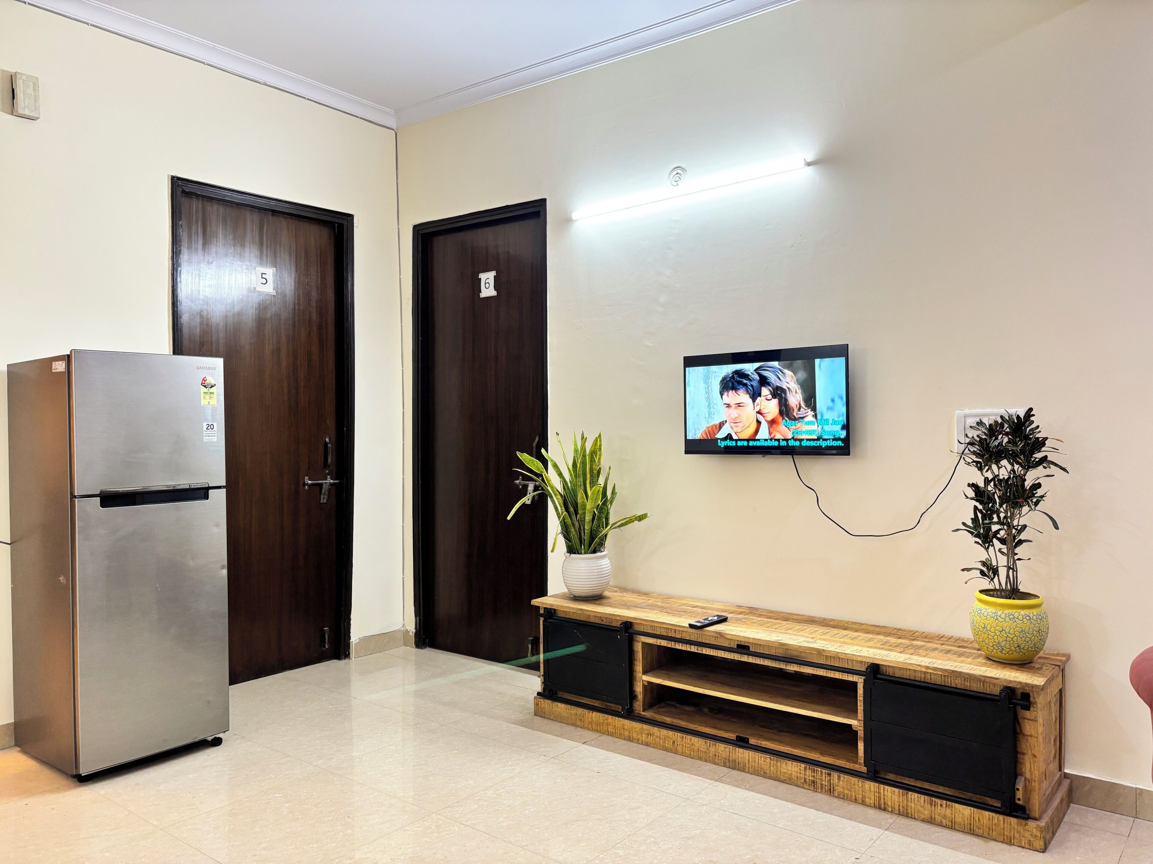 Ein Hotelzimmer mit einem Edelstahlkühlschrank, einer Holztruhe zur Unterhaltungselektronik, einem an der Wand befestigten Fernseher und einer Topfpflanze.