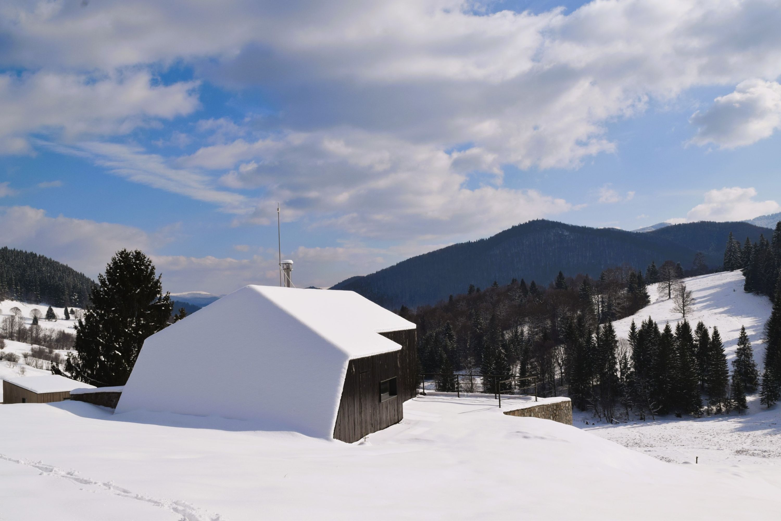 A snow-covered wooden cabin with a sloped roof, surrounded by a snowy landscape and a mountain range in the background.