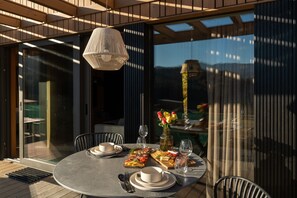 An outdoor dining area with a round table, chairs, a hanging light fixture, and a view through large windows. - Infinity 4p in Vama Buz Ului (Vama Buzăului)
