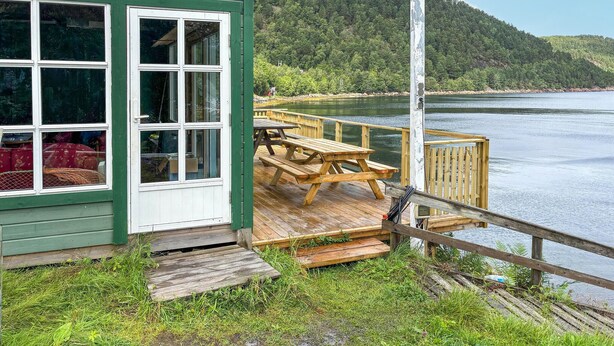A wooden deck with picnic tables overlooking a lake, a green building with large windows, and a forested hillside in the background. - Nice home in Hellandsjøen with WiFi (Hellandsjøen)