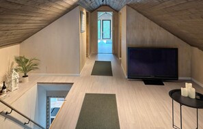 A spacious attic room with a large TV, a small table with candles, and a view through a door to a balcony.
