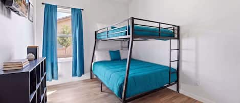 Un lit superposé avec un matelas bleu, un plancher en bois, un ventilateur de plafond et une fenêtre avec des rideaux.